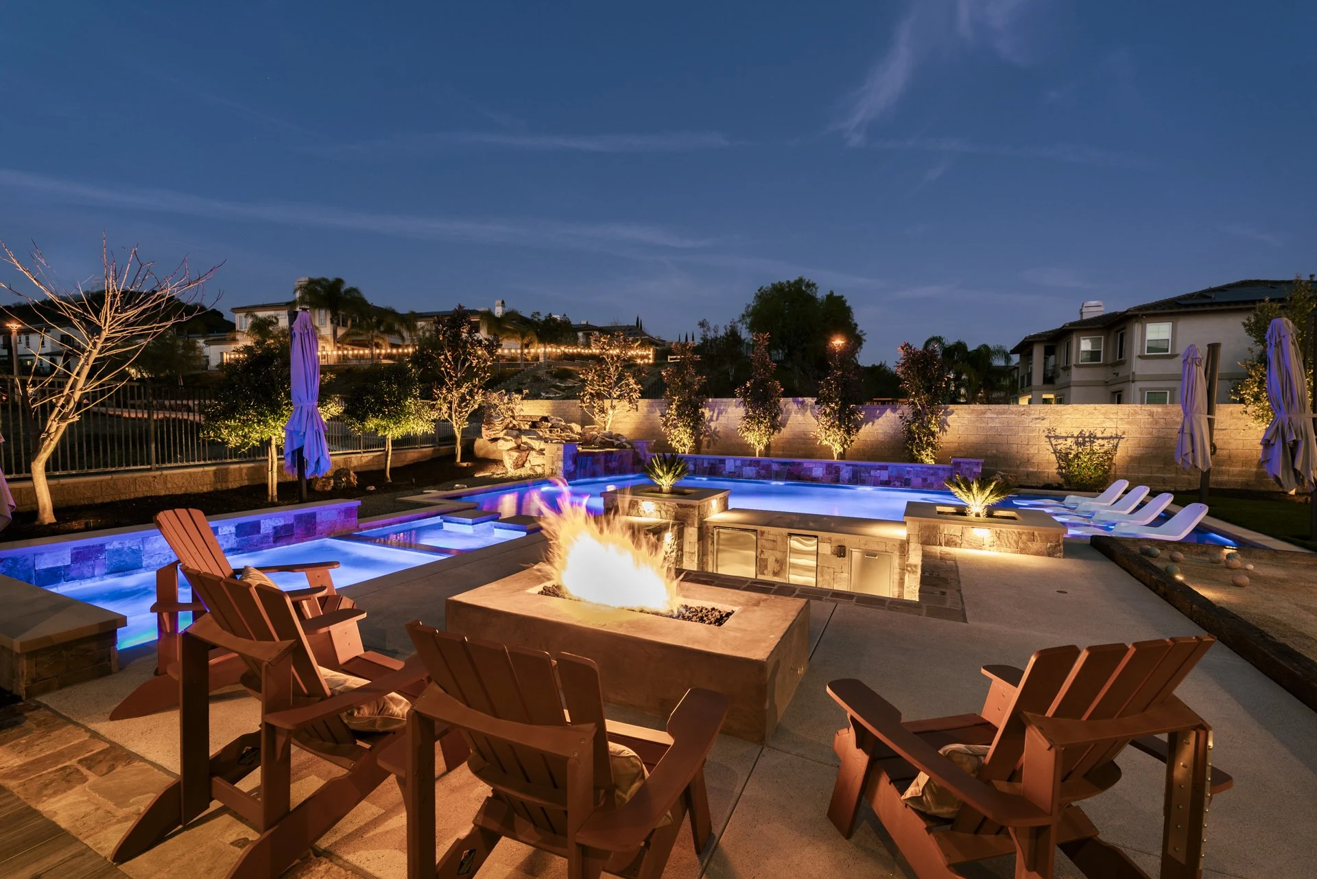 Fire pit and Adirondack chairs at dusk, with a lit pool and spa beyond.
