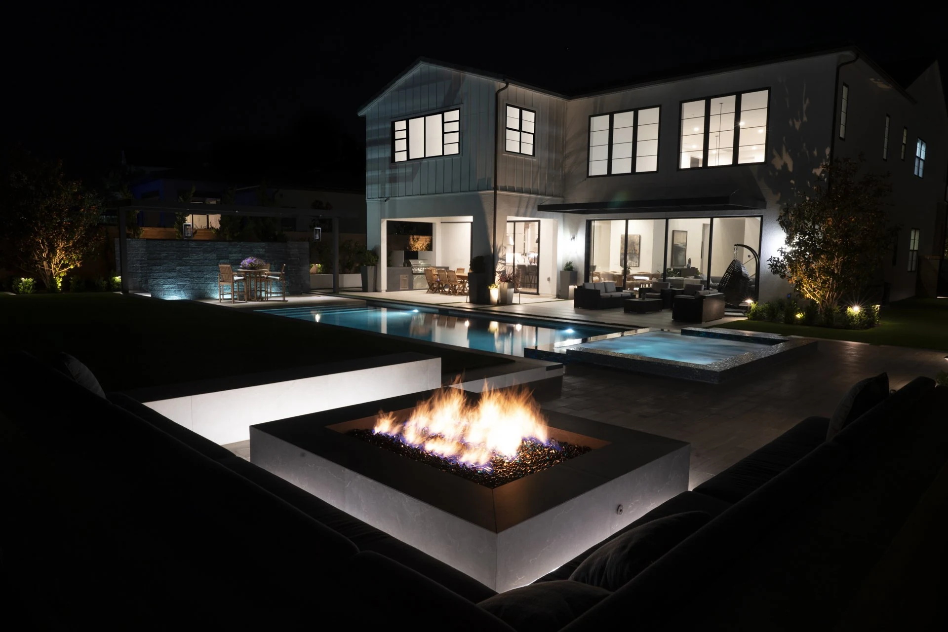 Lit fire pit, pool and spa beside a white house at night.