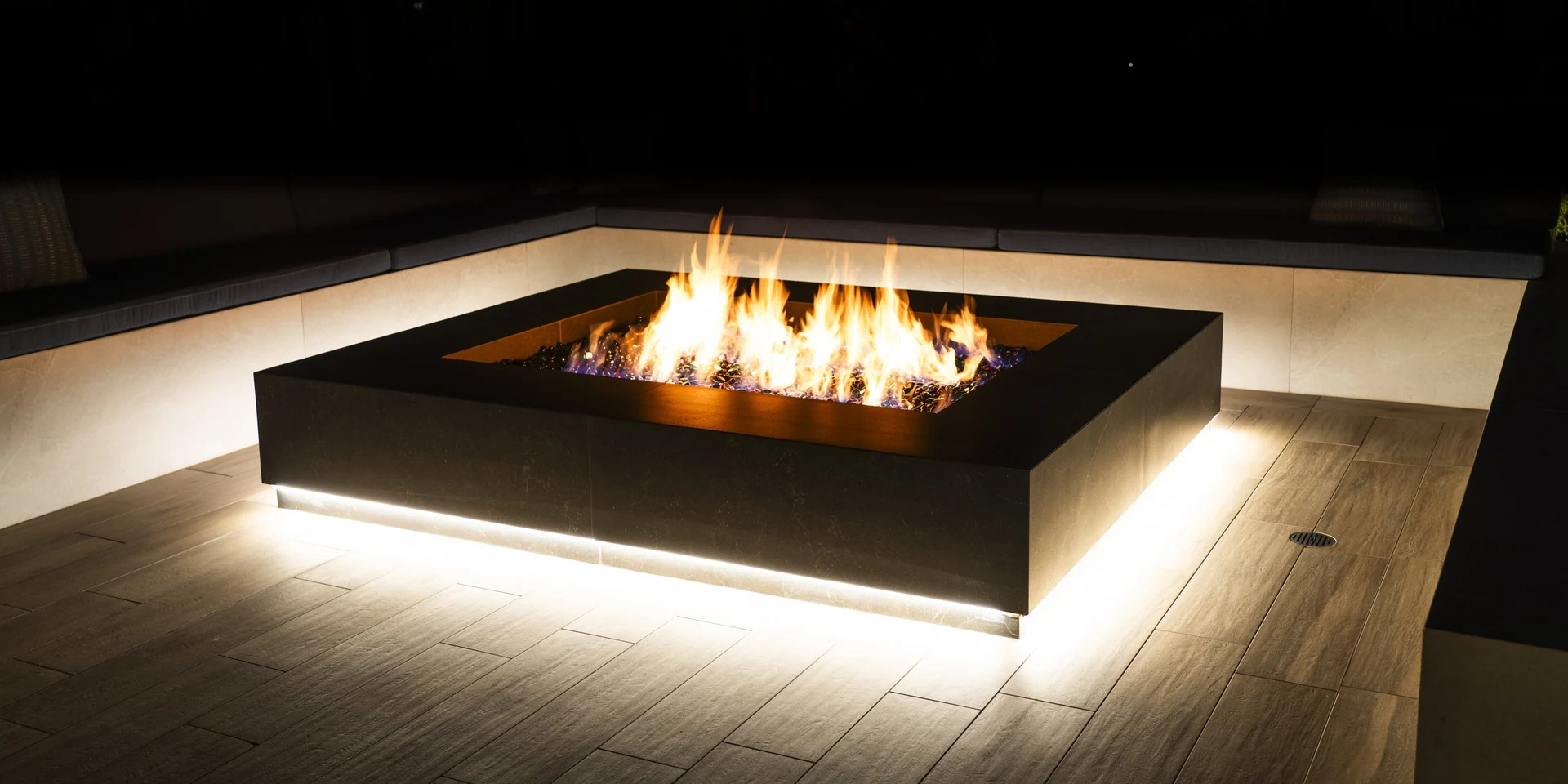 A black fire pit glowing on a wood-look deck at night.